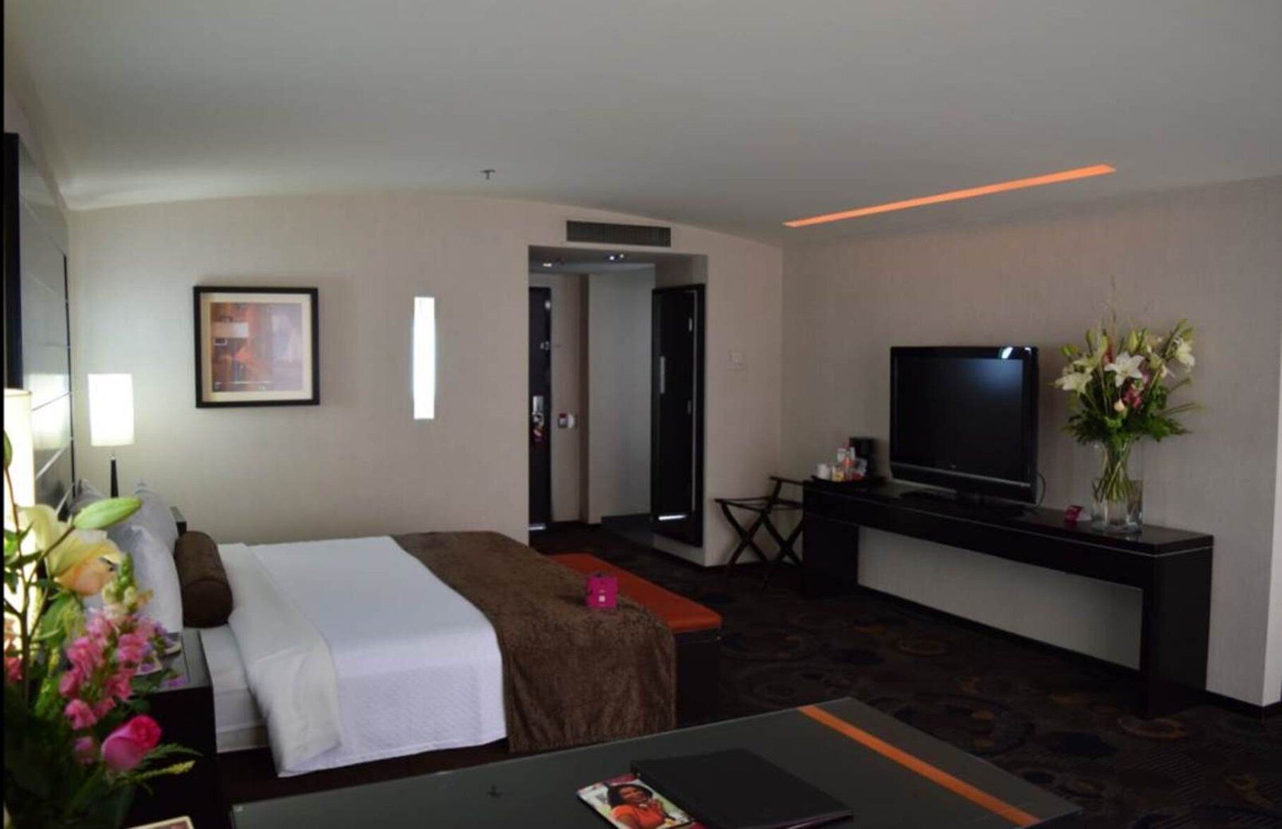 A hotel room with a bed, a desk with a flat-screen TV, a chair, a vase with flowers, and a framed picture on the wall.