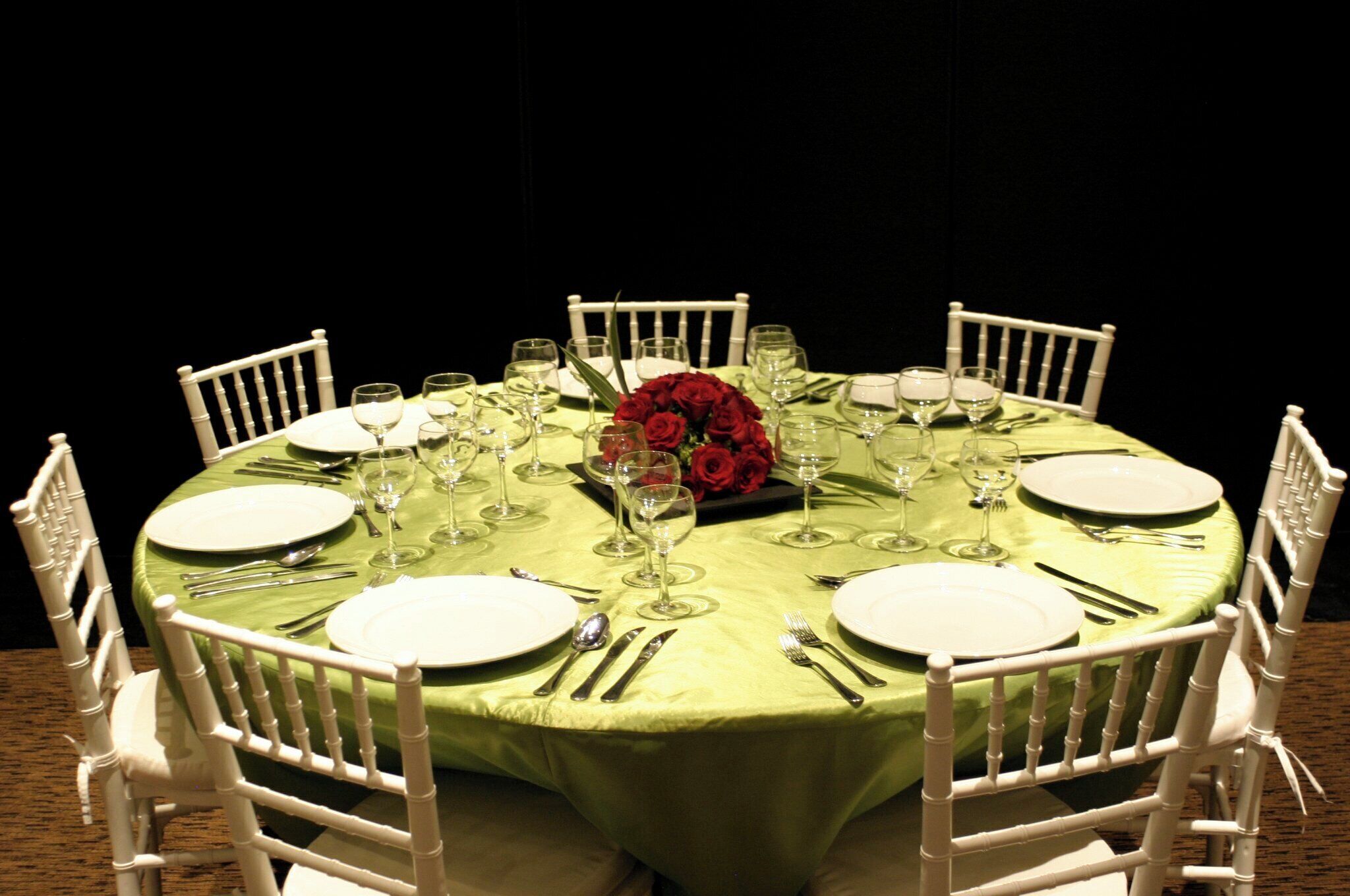 Una mesa redonda dispuesta para una comida, con sillas blancas, mantel verde y un centro de mesa con rosas rojas.