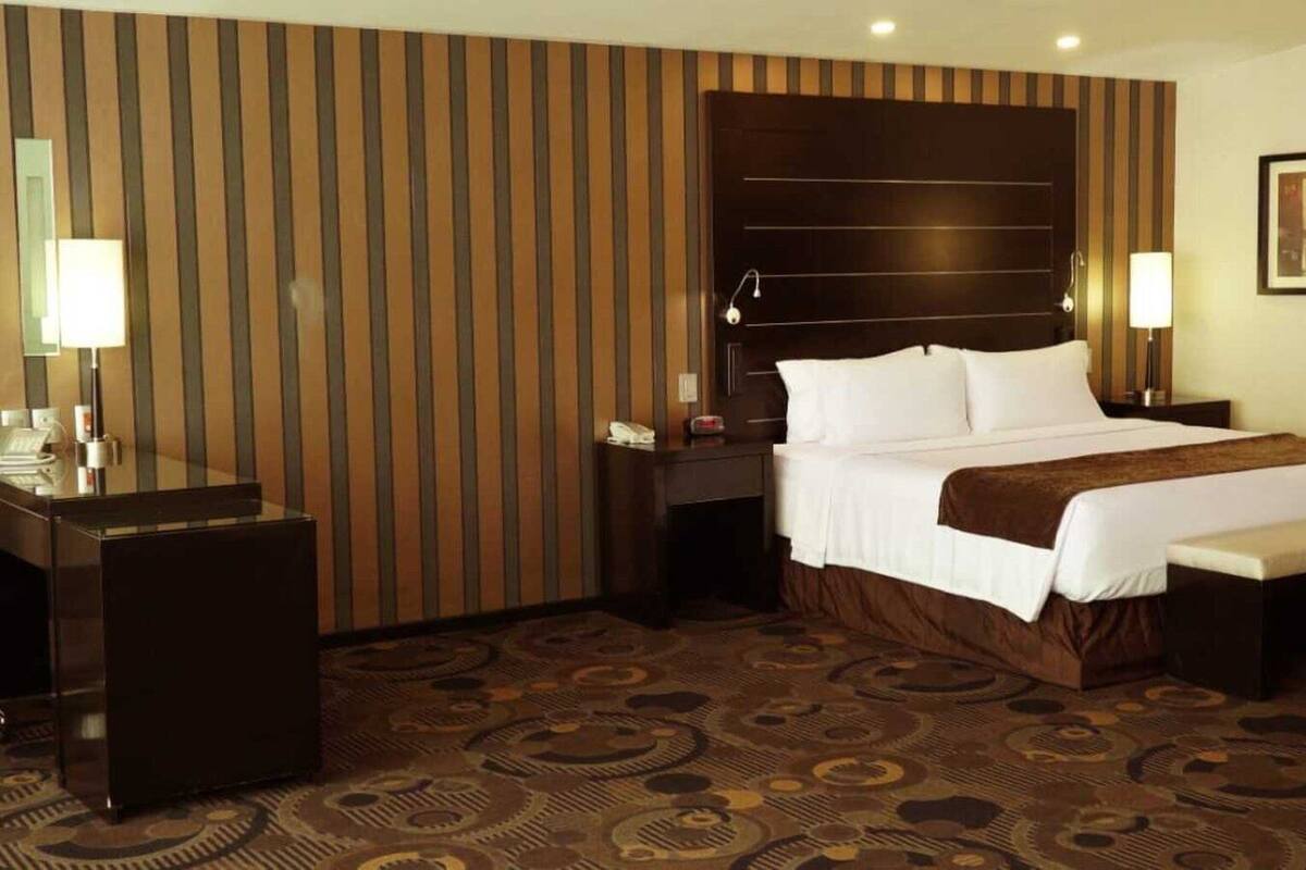 A hotel room with a large bed, a desk, and a chair. There are two bedside tables with lamps. The room has a striped wall and a patterned carpet.