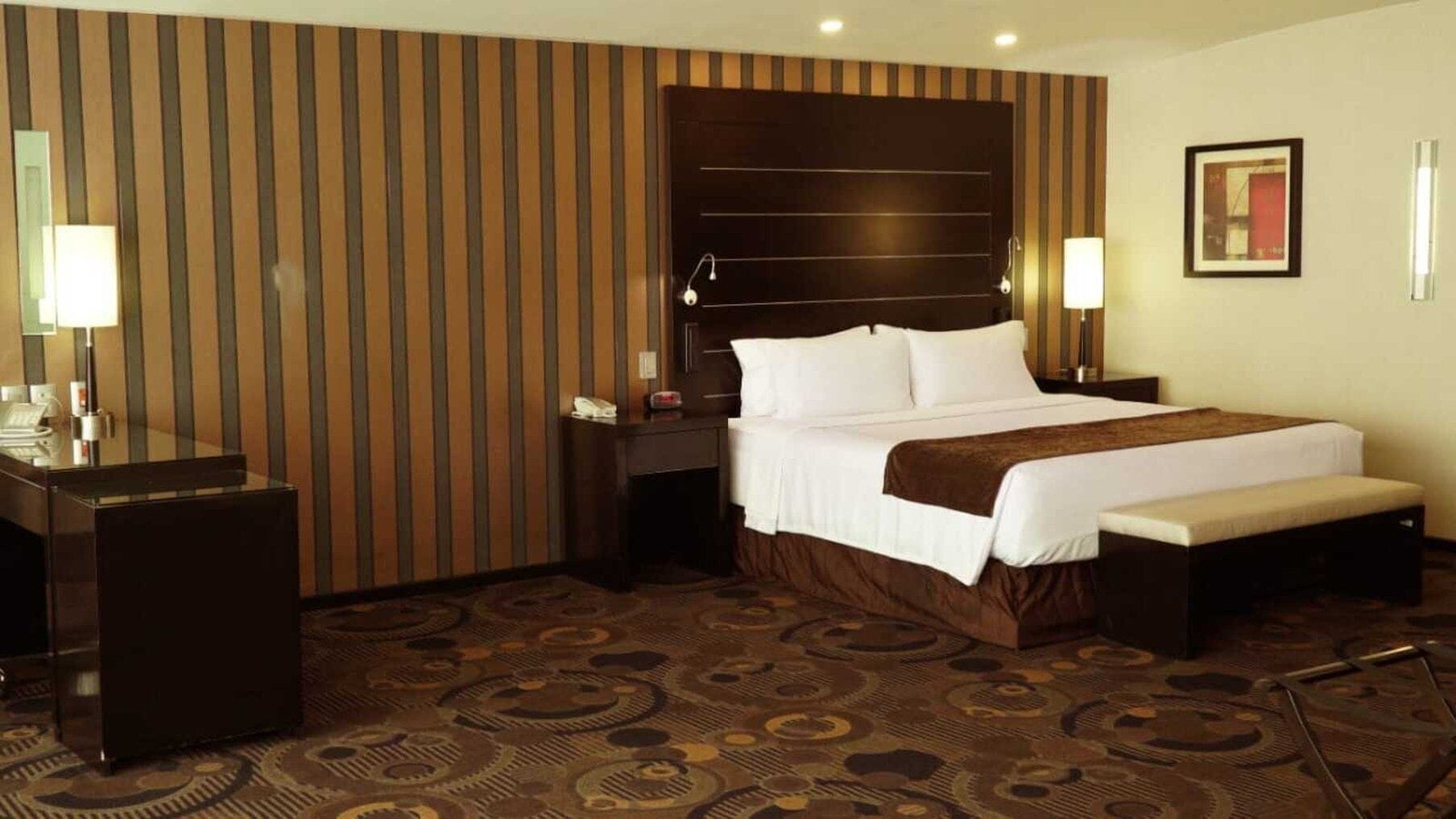 Una habitación de hotel con una cama grande, un escritorio y una silla. Hay dos mesitas de noche con lámparas. La habitación tiene una pared a rayas y una alfombra estampada.