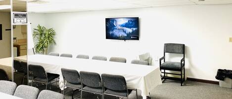 Une salle de conférence avec une longue table, des chaises grises, un téléviseur fixé au mur et une plante en pot.