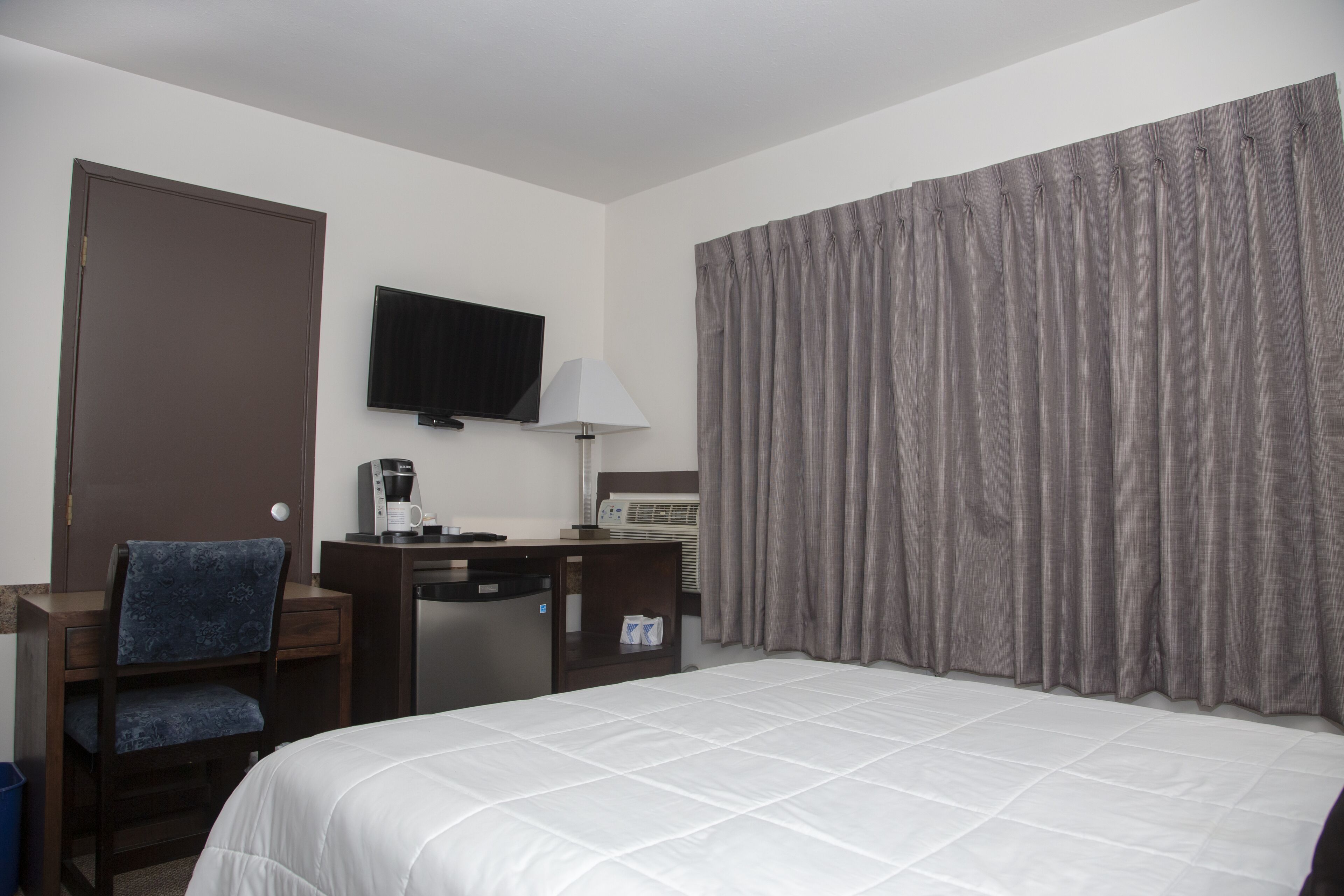 A hotel room with a bed, a desk with a chair, a television, a lamp, and a window with curtains.