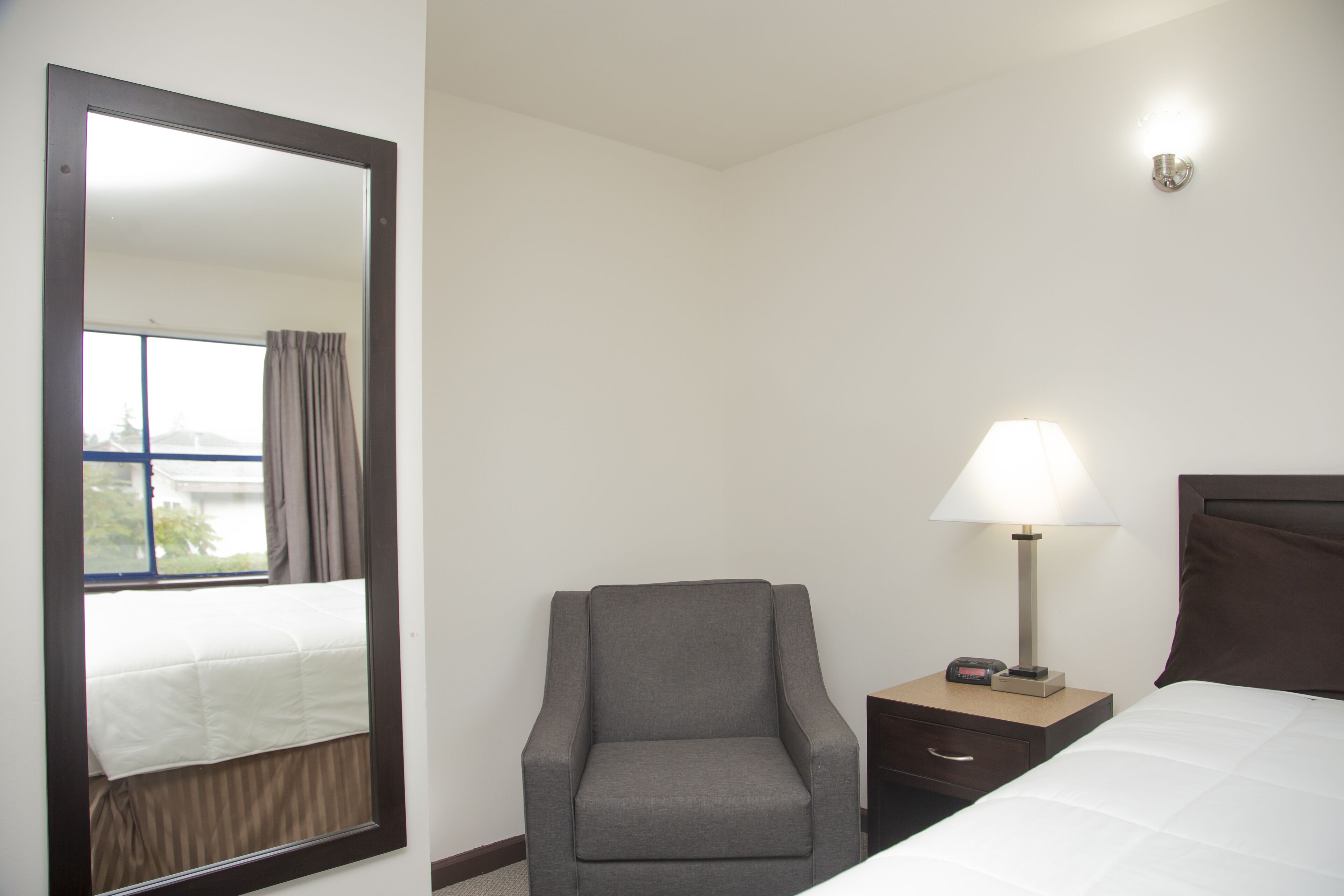 A hotel room with a large mirror, a chair, a bedside table with a lamp, and a bed with a white comforter.