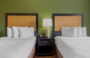 Habitación de hotel con dos camas, cada una con ropa blanca y cabecero. Entre las camas hay una mesita de noche con una lámpara, un teléfono y un reloj. Las paredes son verdes.