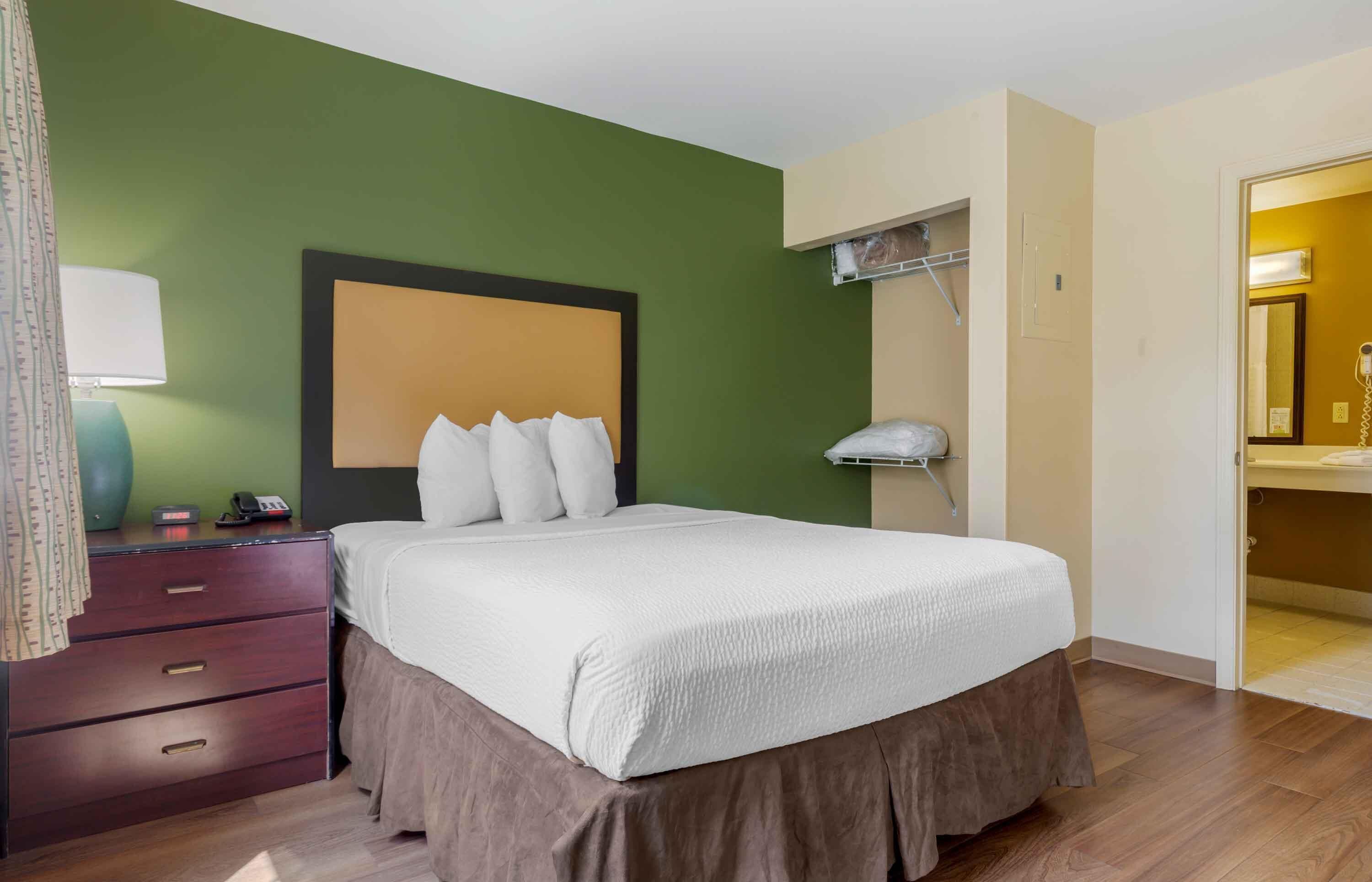 Une chambre d’hôtel avec un lit, une commode, un miroir et une salle de bain visible à travers une porte ouverte.