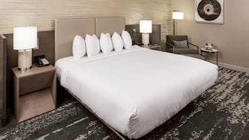 Quarto de hotel com uma cama grande, duas mesas de cabeceira com luminárias, uma cadeira, uma mesinha com luminária e uma obra de arte emoldurada na parede.