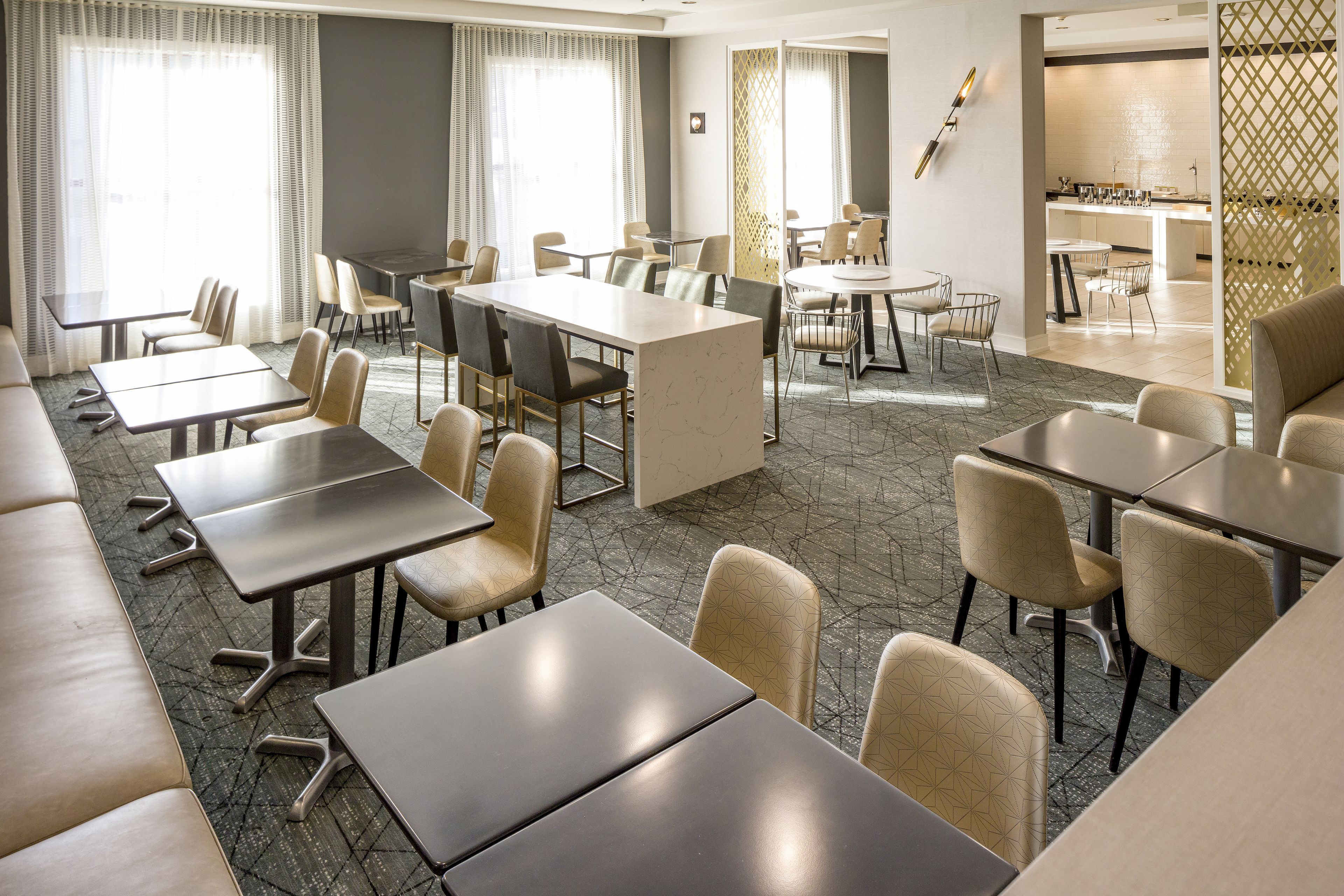 A hotel dining area with multiple tables and chairs, a kitchen in the background, and a view through a partition.