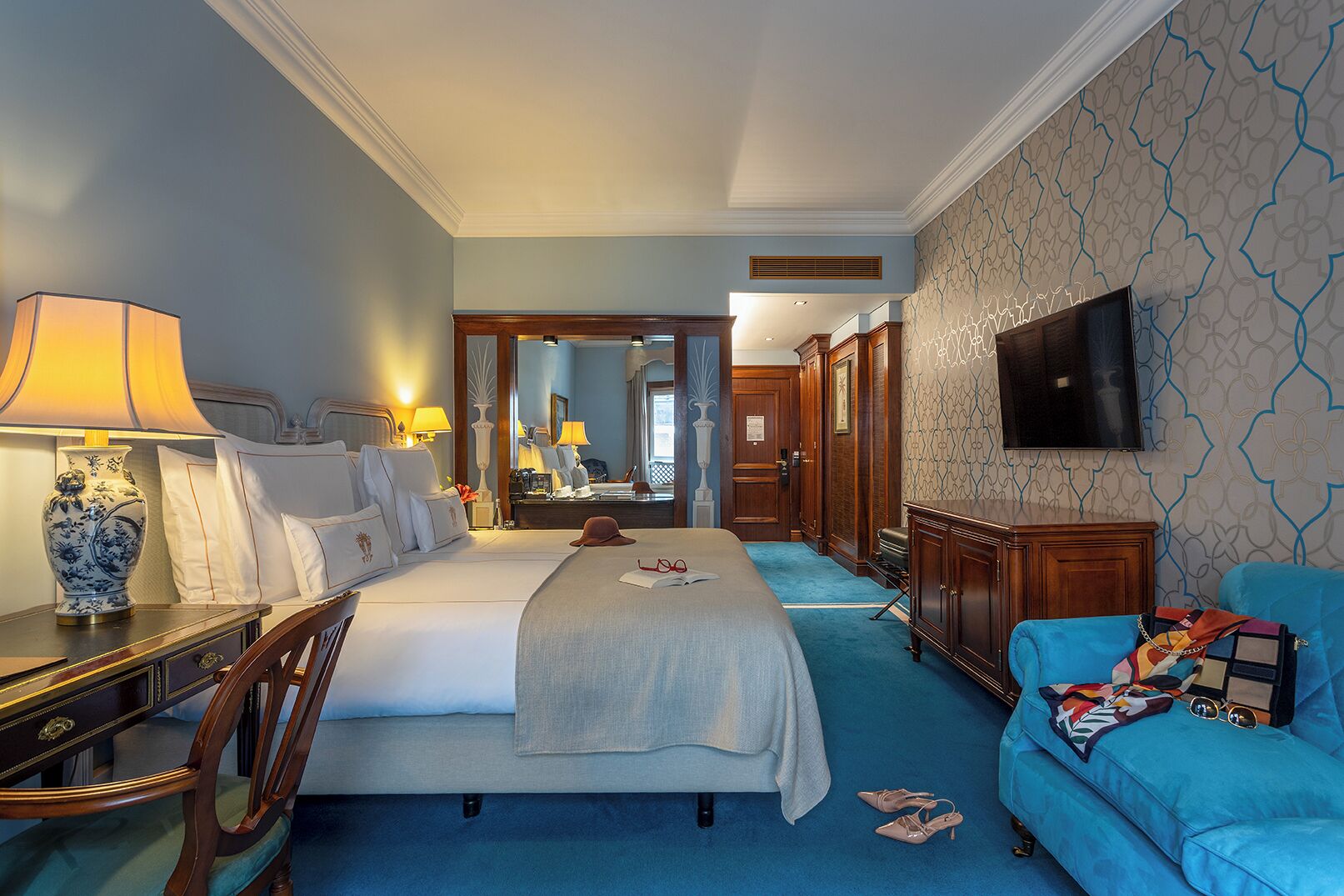 A hotel room with a large bed, a desk with a lamp, a blue sofa, and a television on the wall.