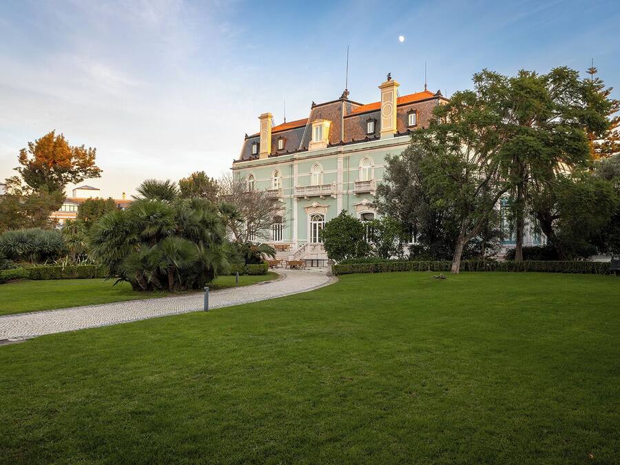 A large, historic building with a central staircase, surrounded by well-maintained lawns and trees.
