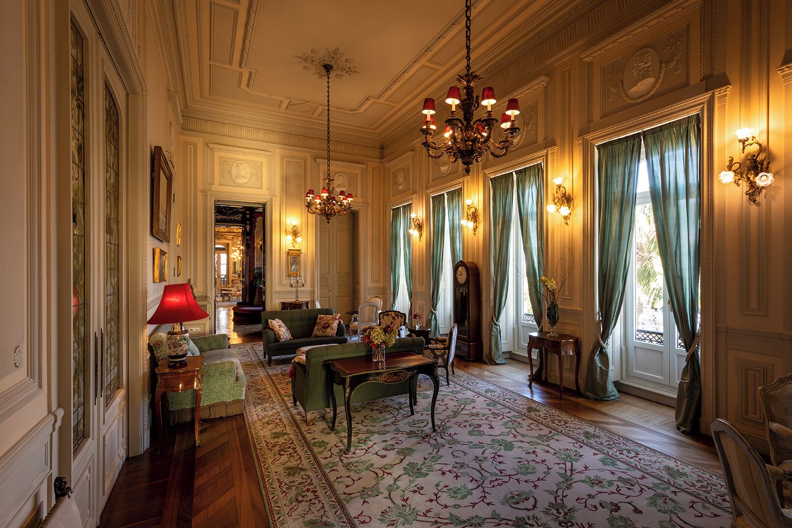 Een statige kamer met sierlijke ornamenten, een kroonluchter, een bank, een tafel met een vaas en een groot raam met gordijnen.