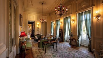 Een statige kamer met sierlijke ornamenten, een kroonluchter, een bank, een tafel met een vaas en een groot raam met gordijnen.