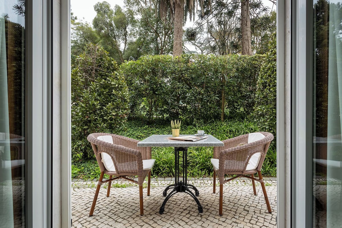 Une terrasse extérieure avec deux fauteuils en osier et une table, entourée d’une végétation luxuriante et d’arbres.