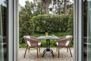 An outdoor patio with two wicker chairs and a table, surrounded by lush greenery and trees.