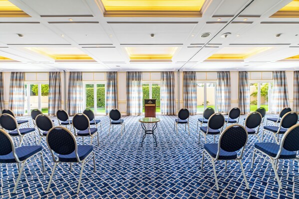 Un salón de conferencias con alfombra estampada, filas de sillas, un atril y ventanas grandes con cortinas.