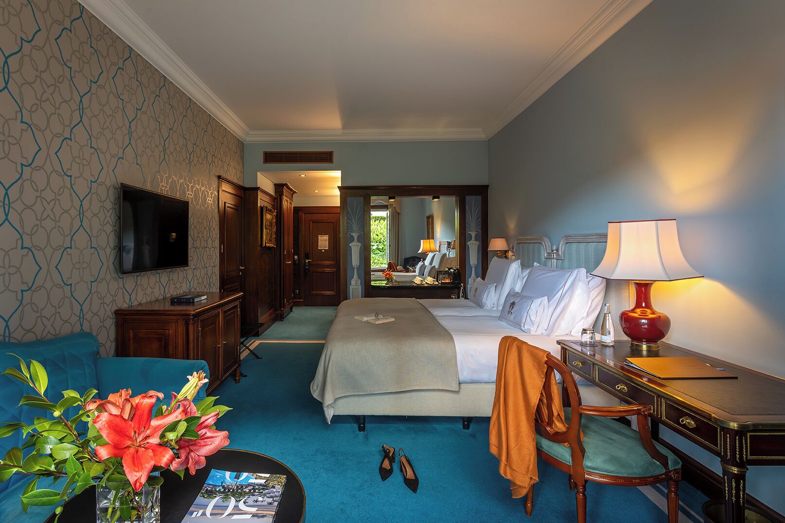 A hotel room with a large bed, a desk with a lamp, a TV, and a view of a garden through the window.