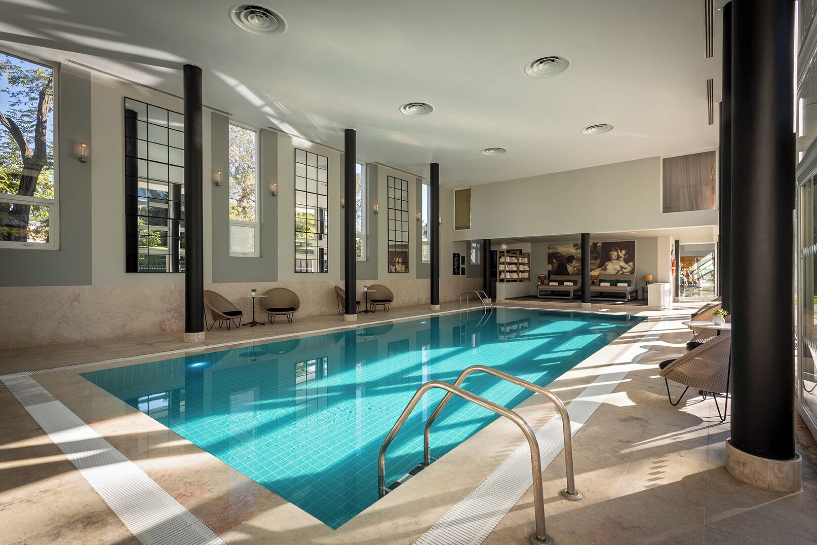 Una amplia piscina interior con agua cristalina, rodeada de sillas de descanso y un espacio luminoso con grandes ventanales.