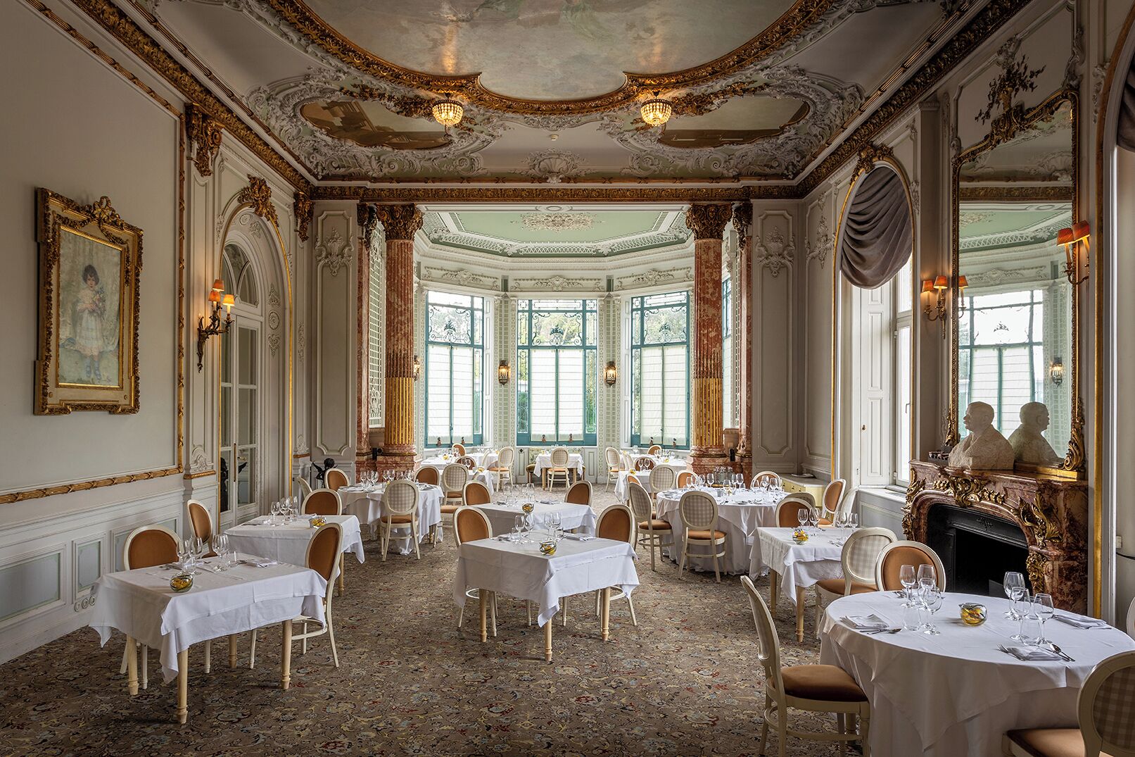 Un comedor amplio y elegante con columnas decorativas, candelabros y una chimenea. Las mesas están dispuestas con manteles blancos y sillas.