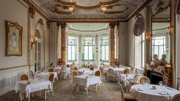 Un comedor amplio y elegante con columnas decorativas, candelabros y una chimenea. Las mesas están dispuestas con manteles blancos y sillas.