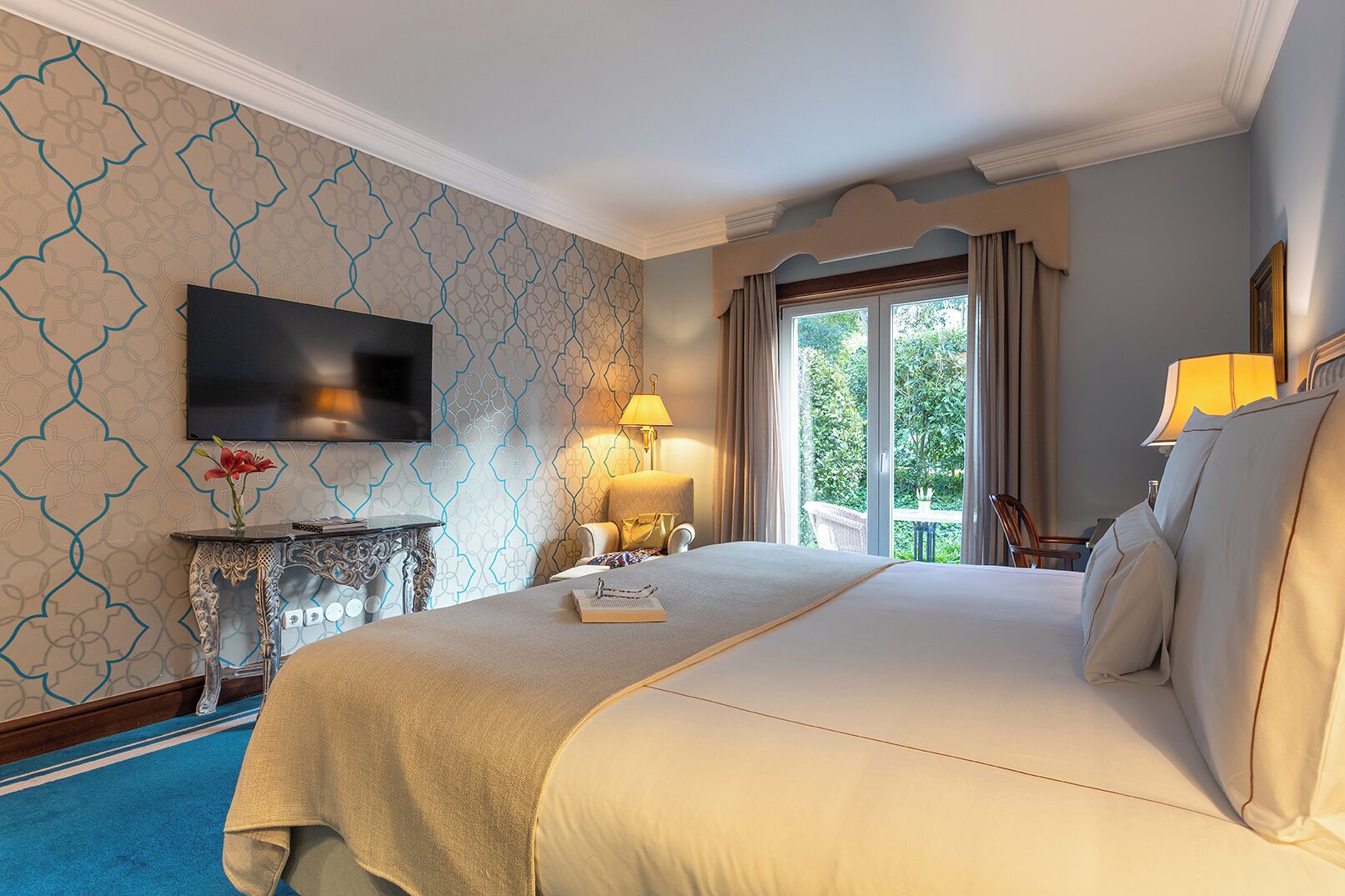 A hotel room with a large bed, a TV mounted on the wall, a bedside table with a lamp, and a view of the outdoors through a window.