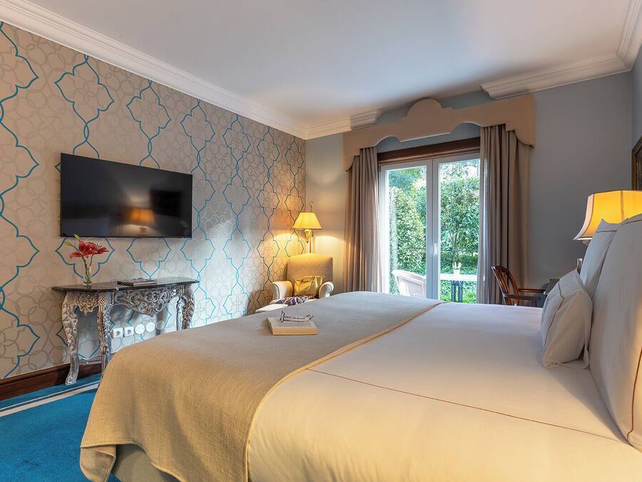 Een hotelkamer met een groot bed, een aan de muur bevestigde televisie, een nachtkastje met een lamp en uitzicht op het omliggende landschap door een raam.