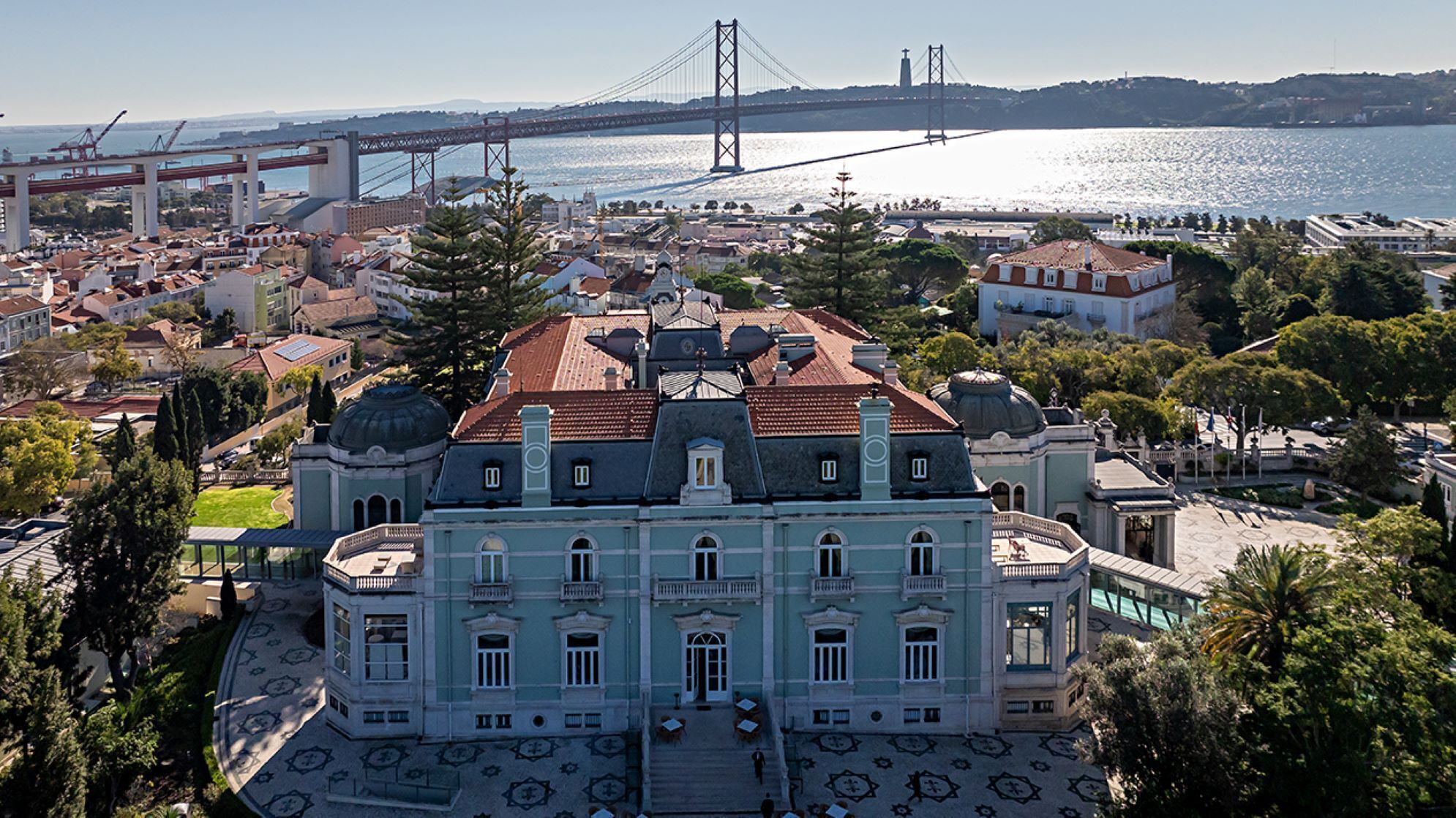 A large, historic building with a central courtyard, surrounded by trees and a view of a bridge over water.