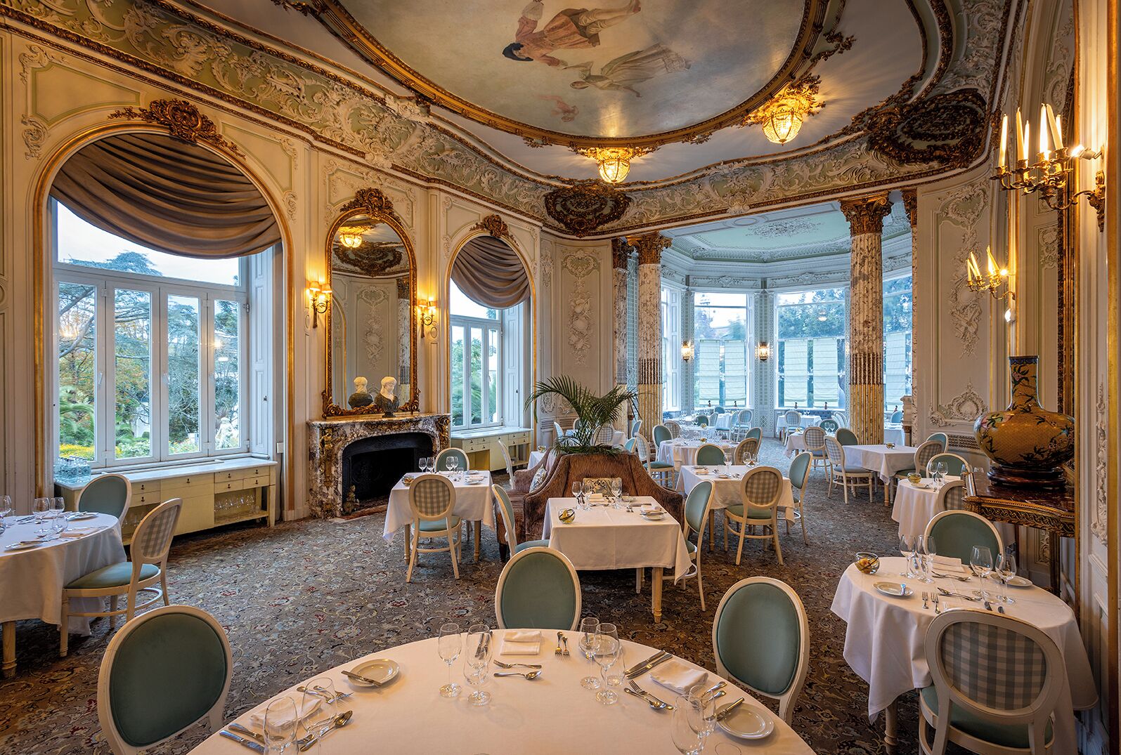 A grand dining room with ornate decor, large windows, and multiple dining tables set for guests.