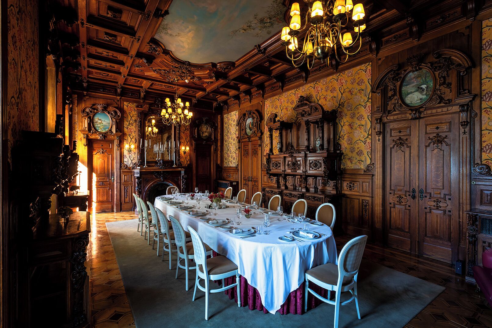 A formal dining room with a long table set for a meal, surrounded by ornate wooden paneling and a large chandelier.