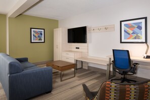 Una habitación de hotel moderna con sofá, escritorio con ordenador, televisor de pantalla plana y dos cuadros en la pared.