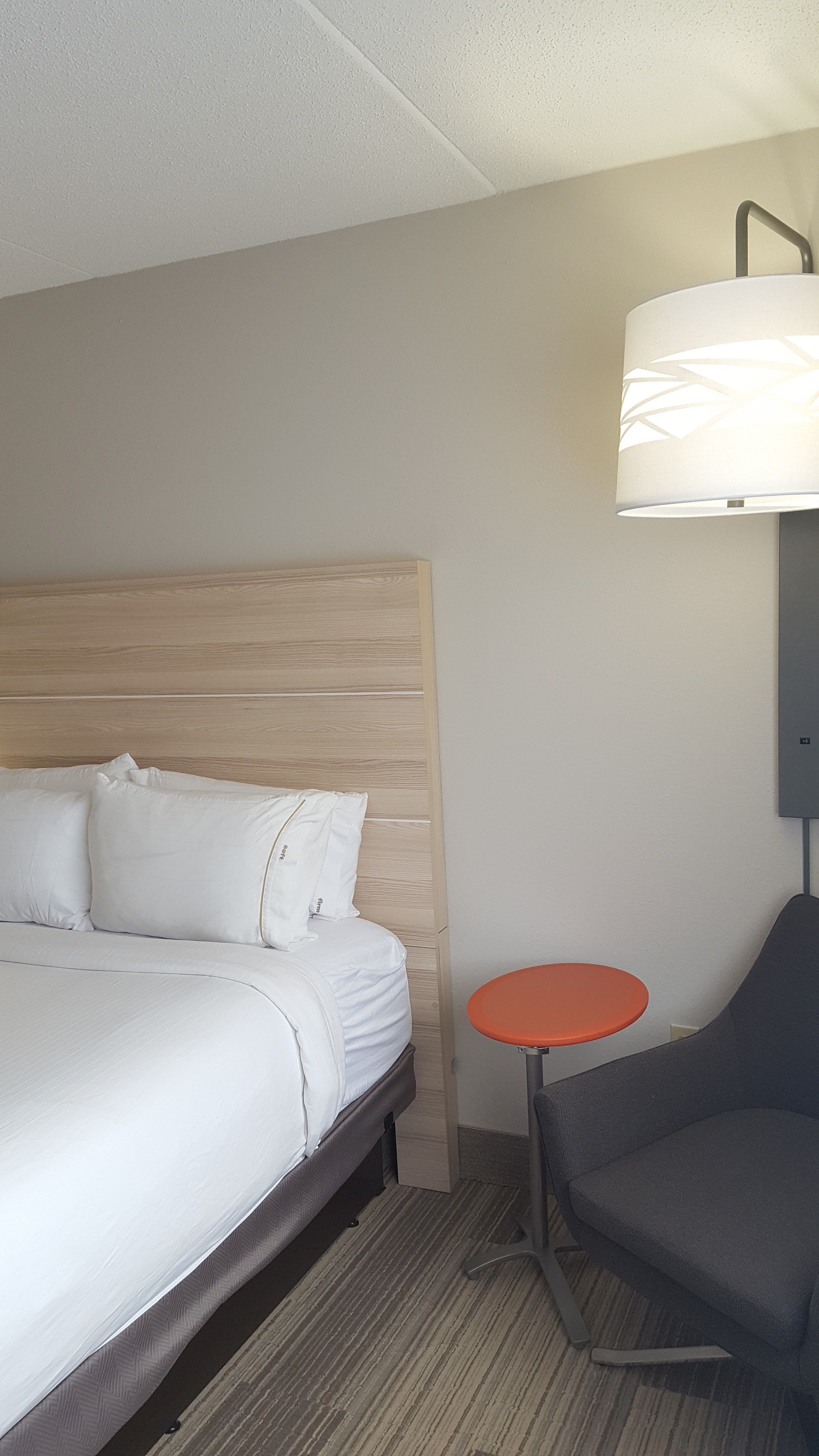 A hotel room with a bed, a small round table, a chair, and a wall-mounted lamp.
