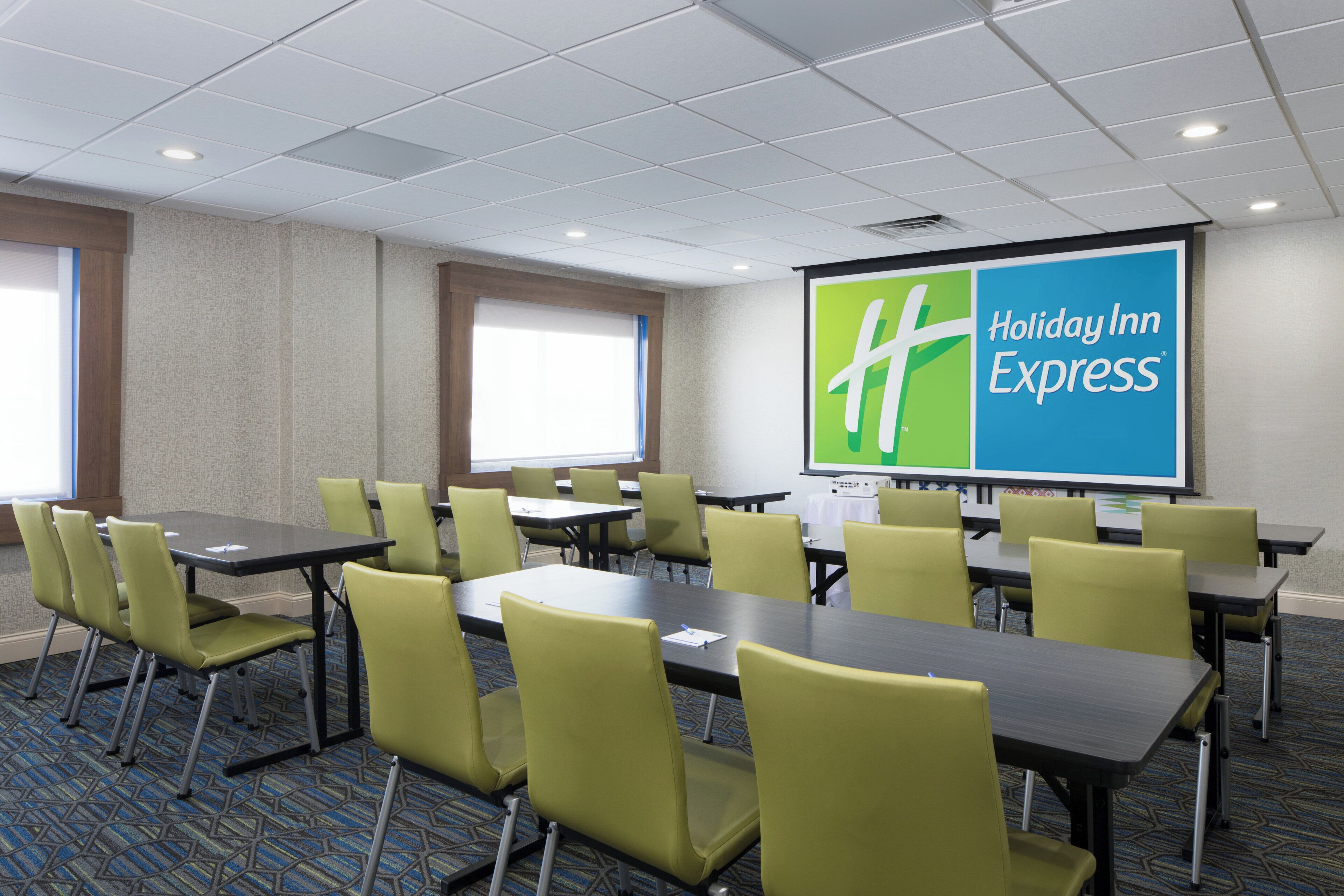 A conference room with tables and chairs, a projector screen displaying 'Holiday Inn Express' branding, and a window providing natural light.