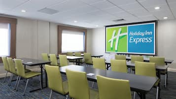 A conference room with tables and chairs, a projector screen displaying 'Holiday Inn Express' branding, and a window providing natural light.