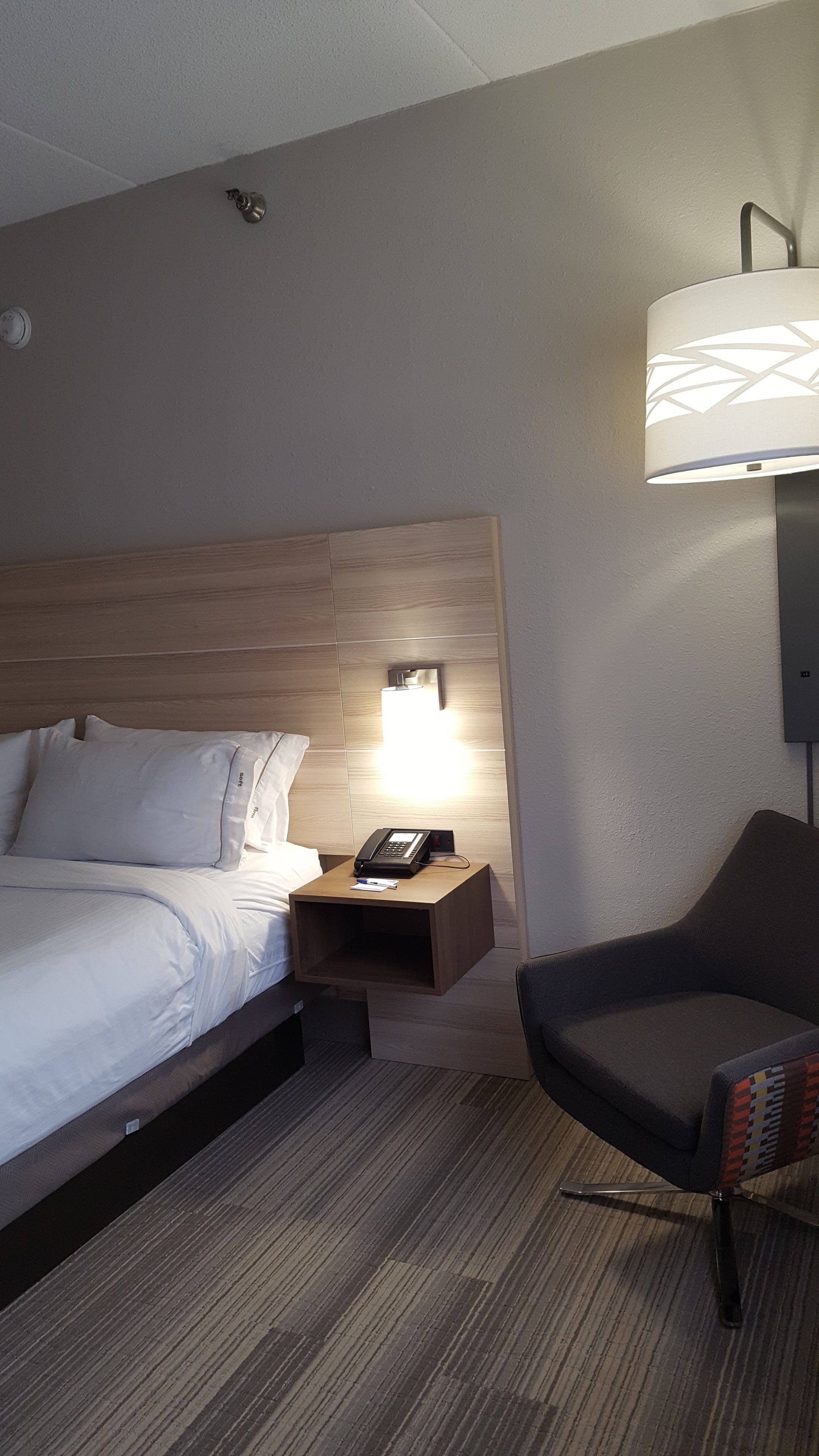 A hotel room with a bed, bedside table with a phone, a chair, and a wall-mounted lamp.