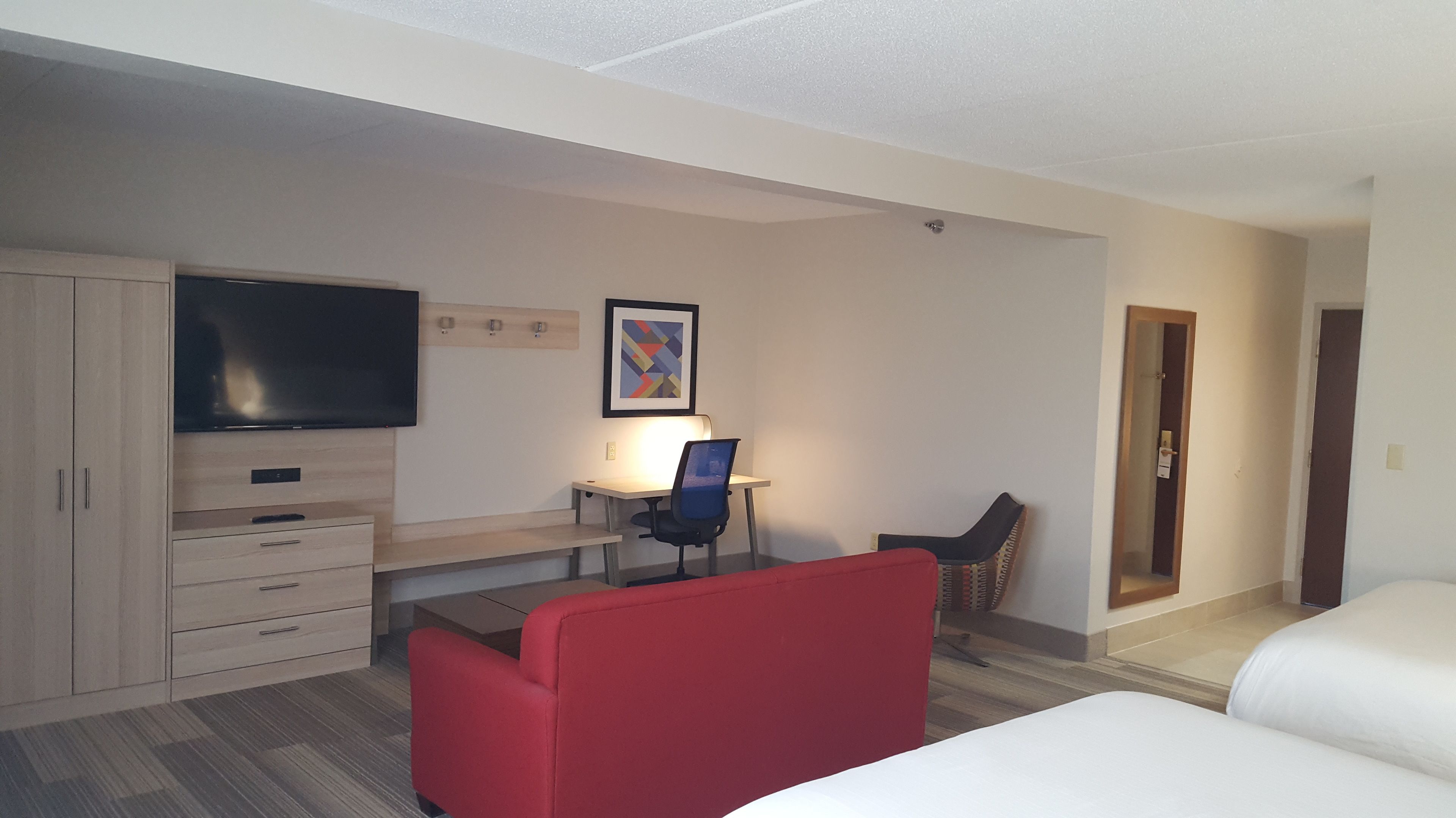 A hotel room with a bed, a desk with a chair, a television, a framed picture on the wall, and a wardrobe.