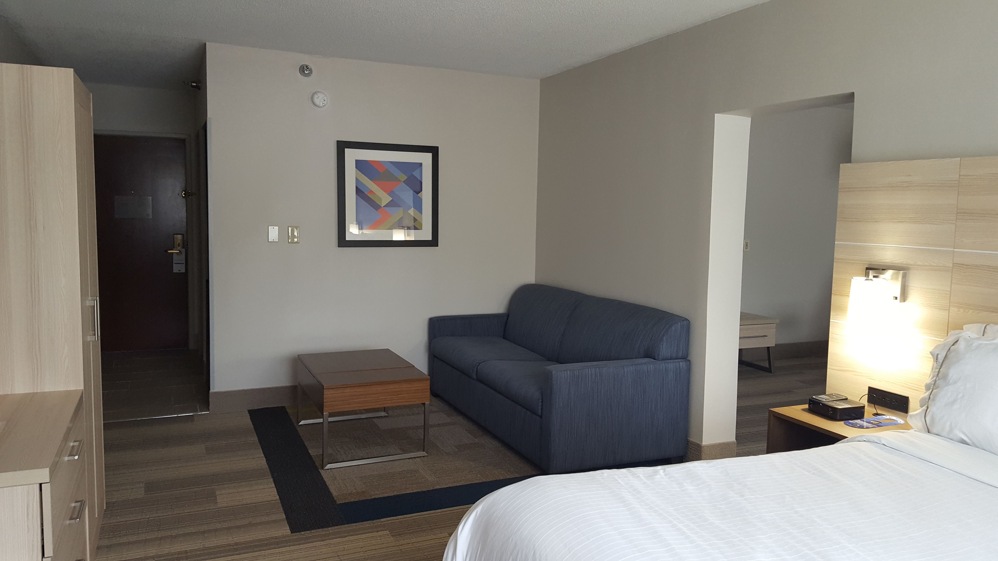 A hotel room with a bed, a blue sofa, a small wooden side table, a framed abstract artwork on the wall, and a nightstand with a lamp.