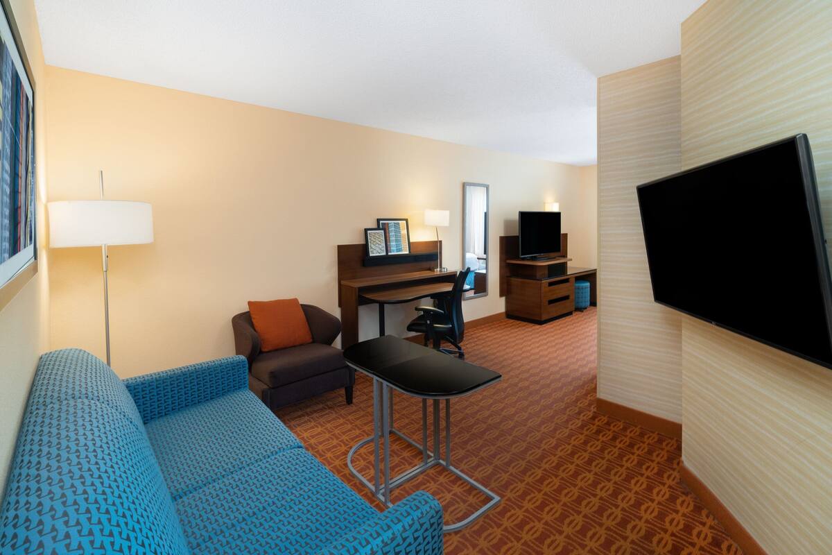 A hotel room with a blue patterned sofa, a small desk with a chair, a television, and a lamp.