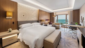 A modern hotel room with a large bed, a desk, a chair, and a view of the ocean.