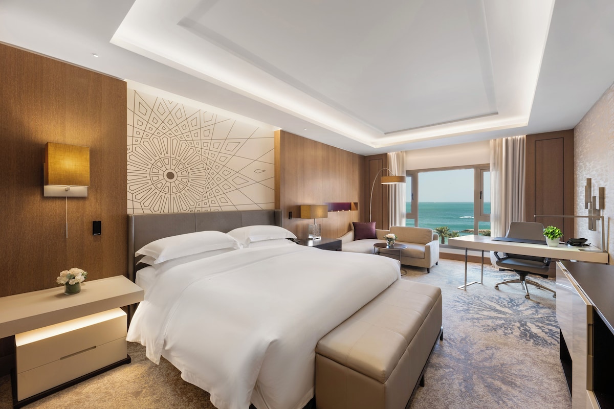 A modern hotel room with a large bed, a desk, a chair, and a view of the ocean.