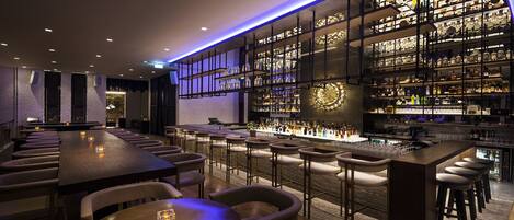 Una zona de bar con una barra larga, varios taburetes y estantes llenos de botellas.
