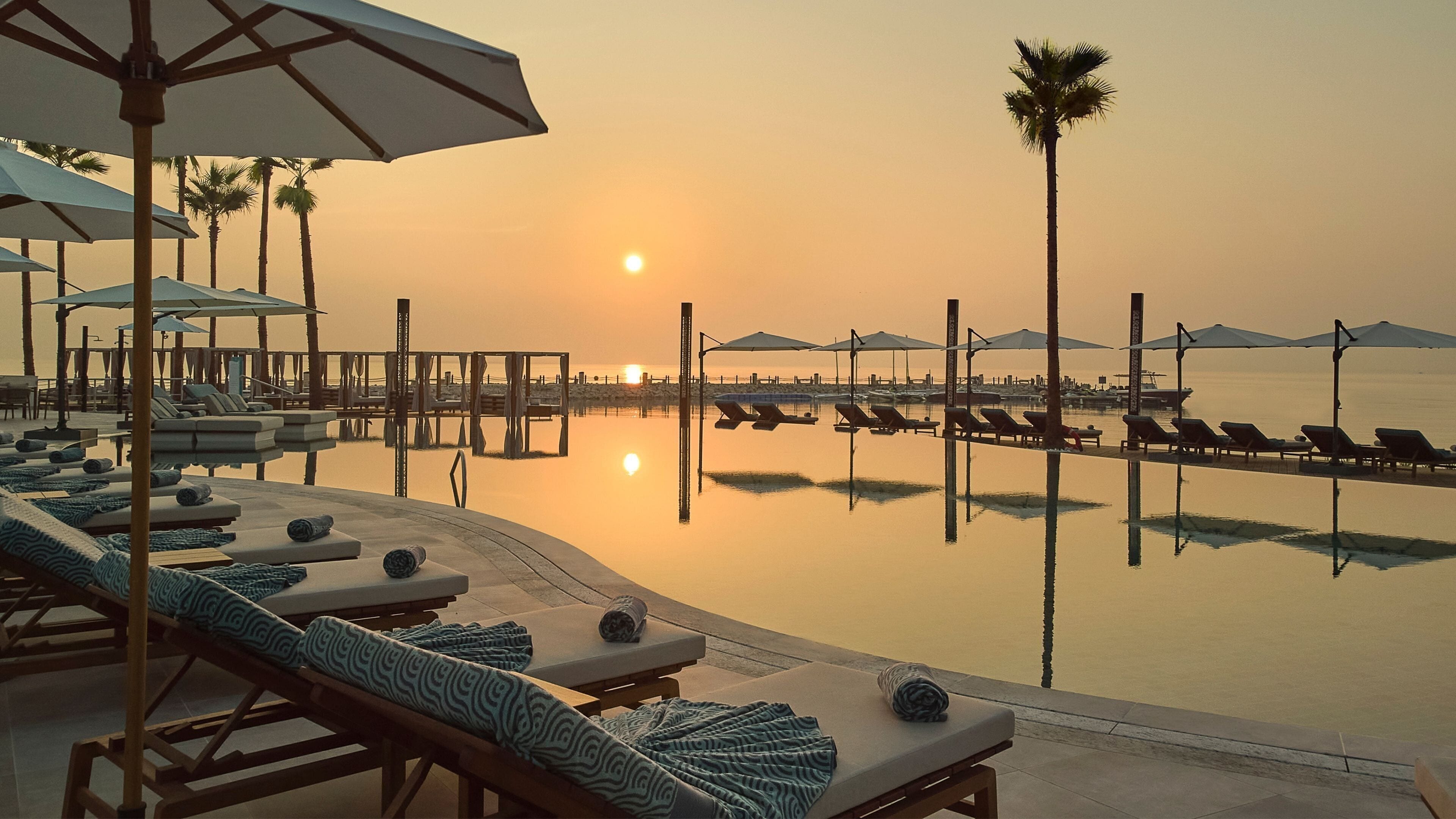 A serene poolside area with lounge chairs and umbrellas, a calm water body reflecting the sunset, and palm trees.