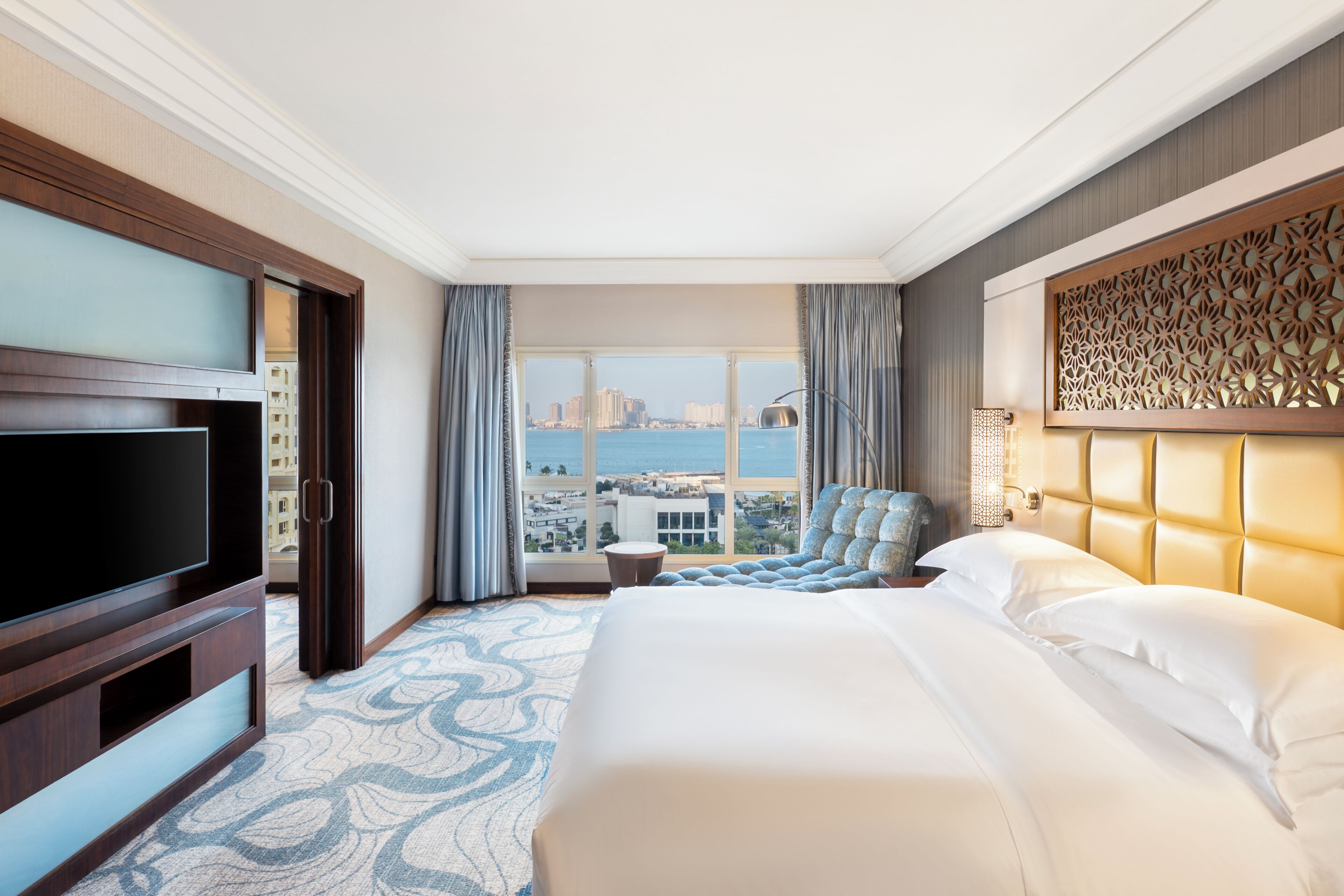 A hotel room with a large bed, a TV, a view of the city, and a decorative wall panel.