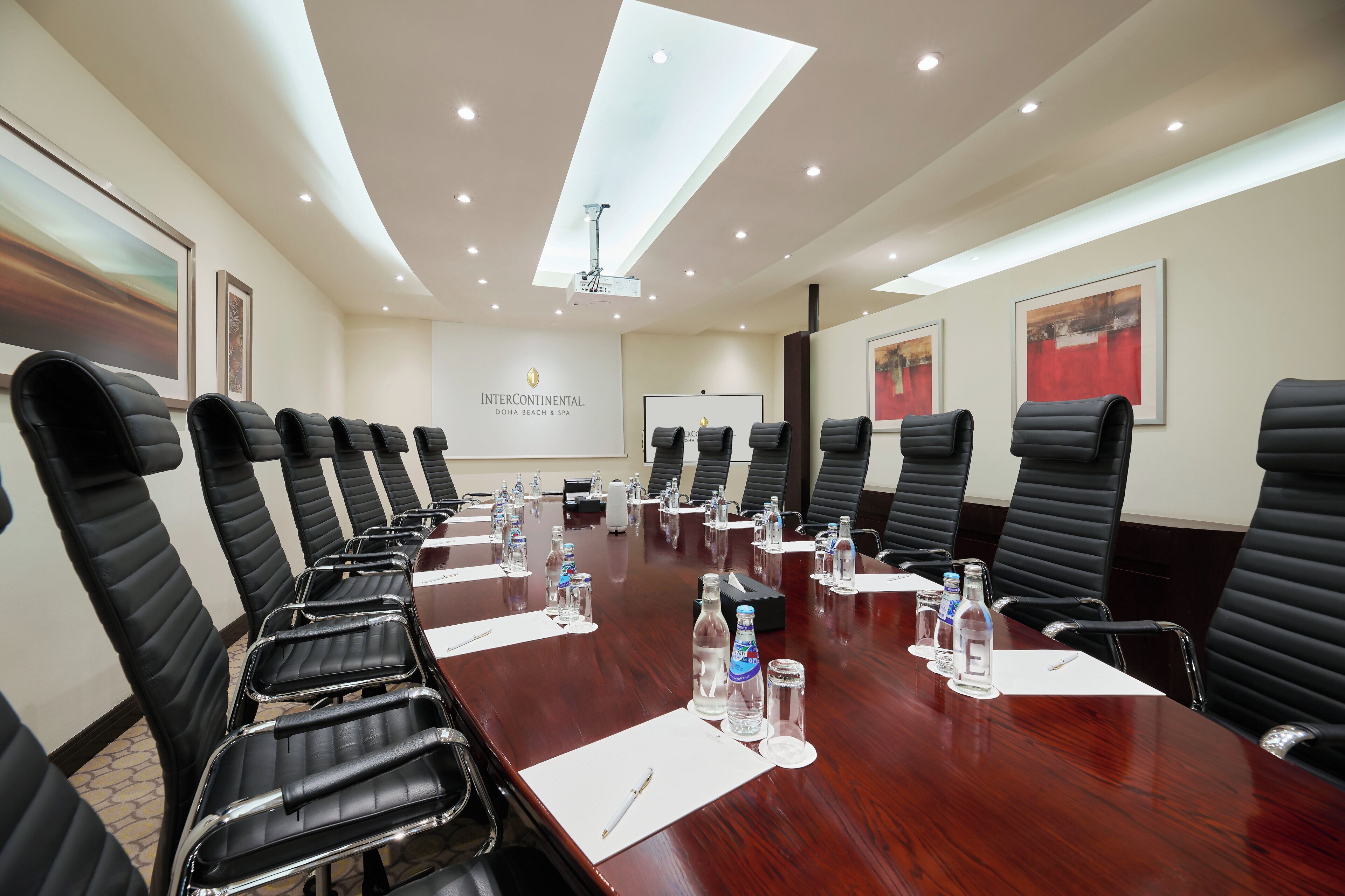 A conference room with a long table, black leather chairs, water bottles, and a projector screen.