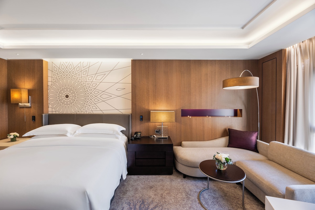A hotel room with a bed, a sofa, a nightstand, a lamp, and a decorative wall piece.
