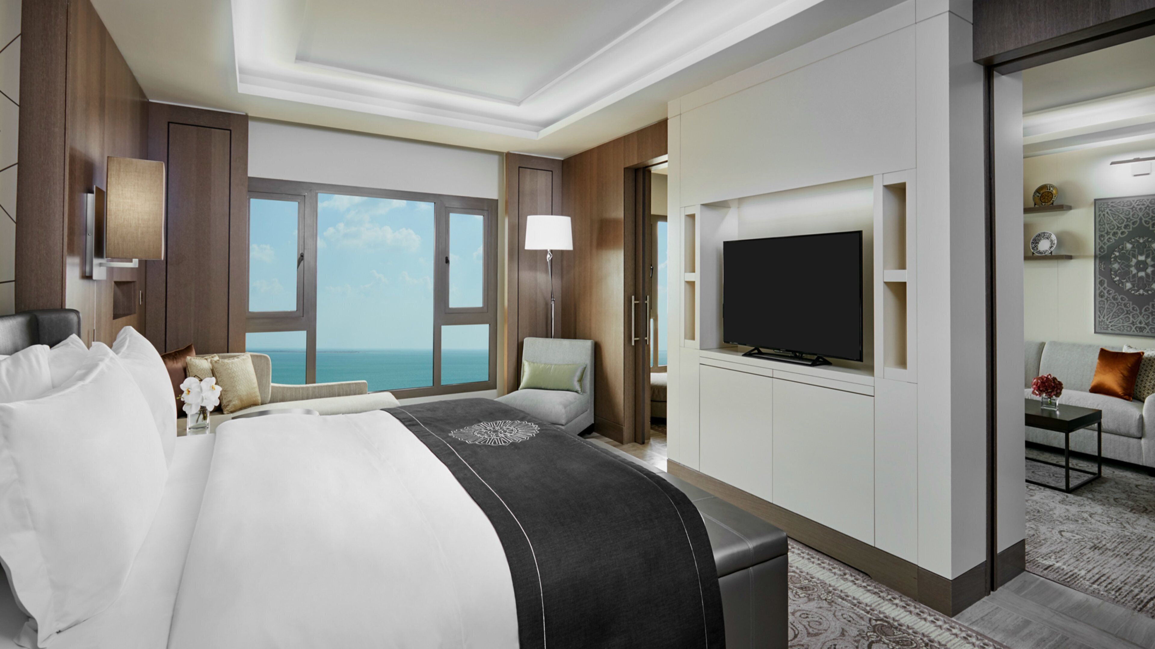 Habitación de hotel con una cama grande, televisión, zona de estar con sofá y vistas al océano a través de amplios ventanales.