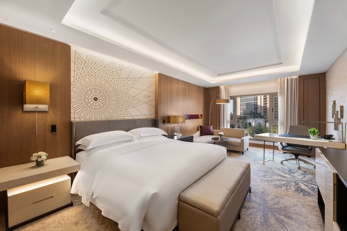 A hotel room with a large bed, a desk, a chair, a sofa, and a view of the city.