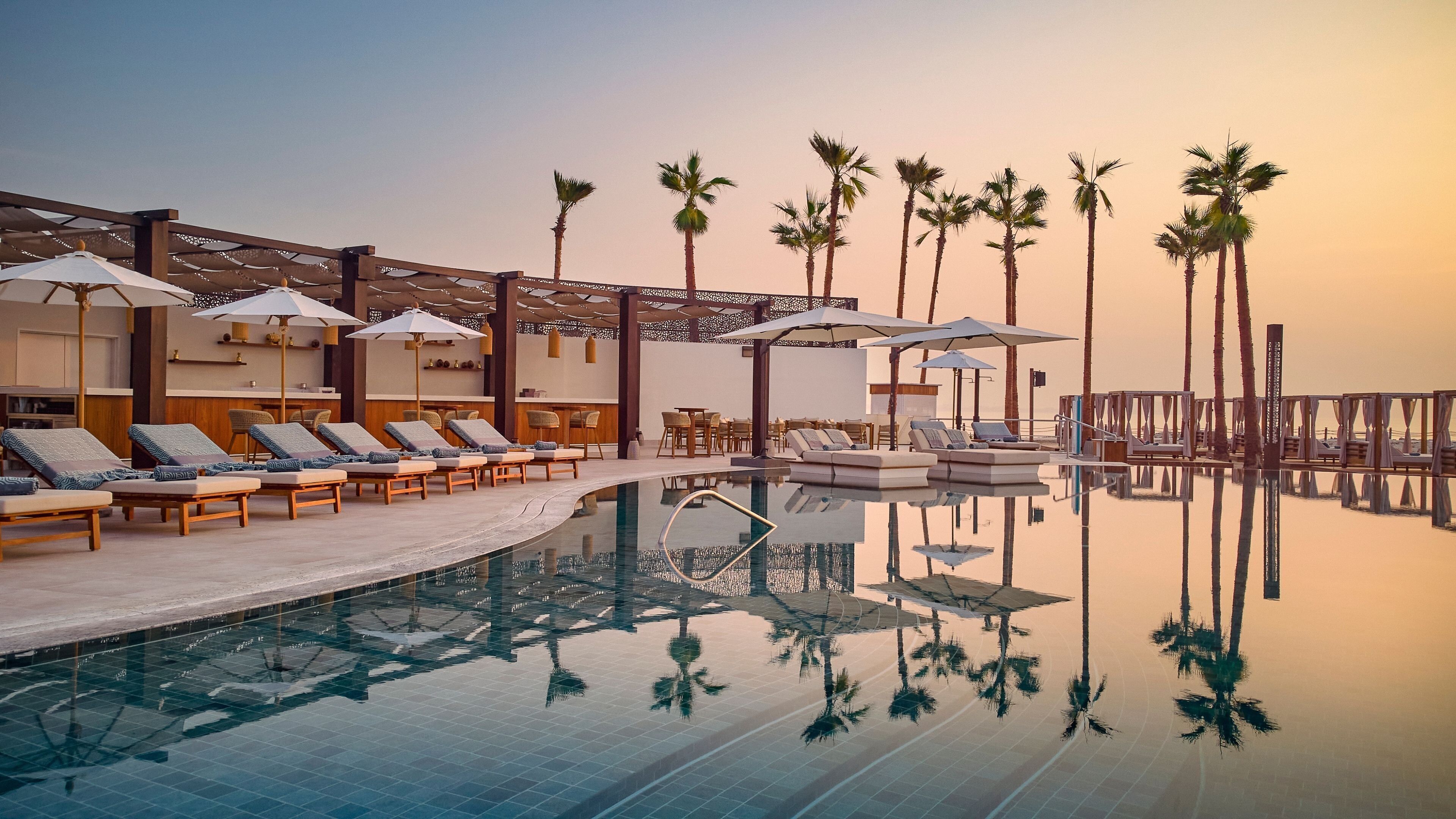 Un espace aménagé au bord de la piscine, avec des chaises longues, des parasols et des palmiers.