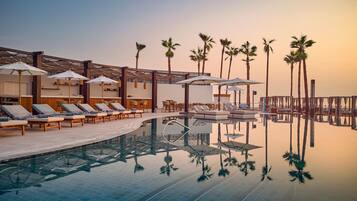 Zona de piscina con sillones reclinables, sombrillas y palmeras.