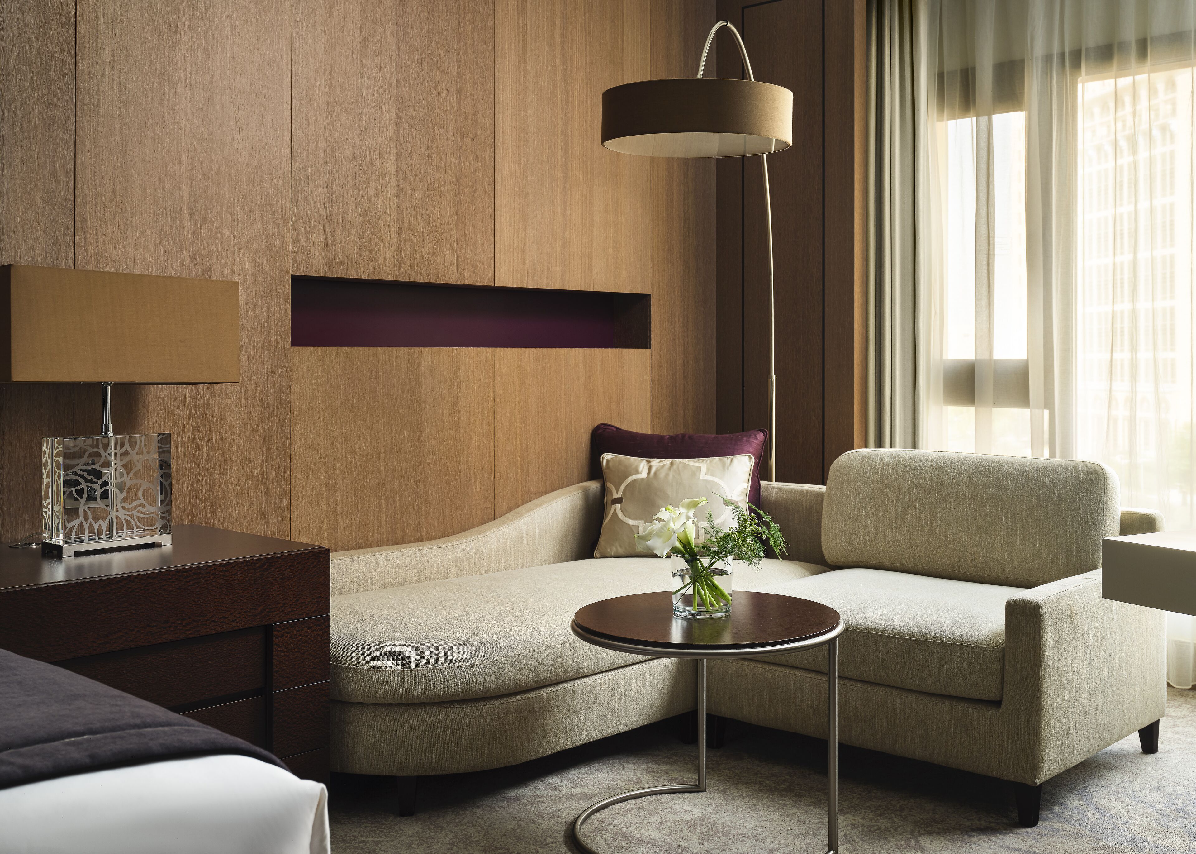 A modern hotel room with a curved sofa, a small round table with a vase of flowers, a lamp, and a view of the city through sheer curtains.