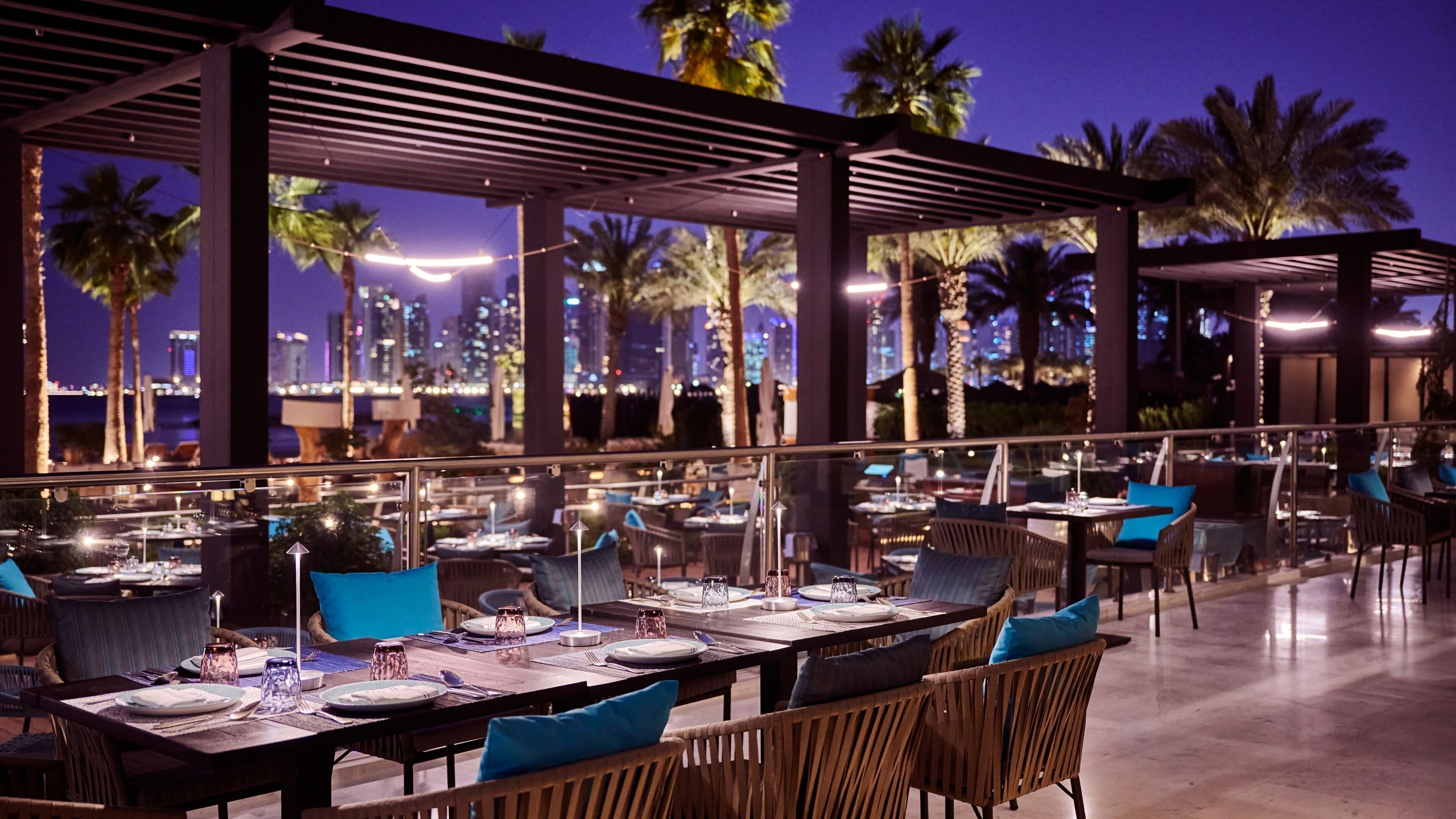 An outdoor dining area with tables and chairs, palm trees, and a cityscape view at night.