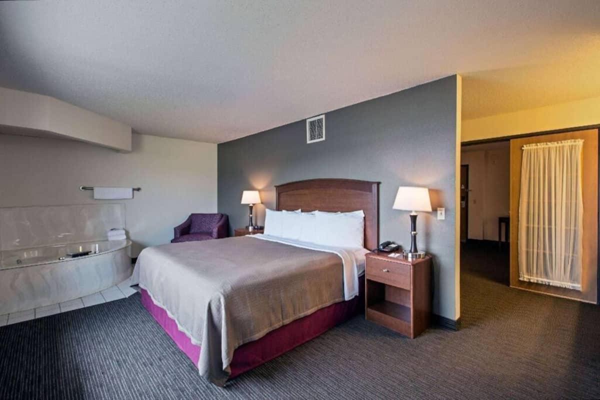 Ein Hotelzimmer mit einem Bett, zwei Nachttischlampen, einem Nachttisch, einem Sessel, einer Badewanne und einer Tür zu einem anderen Raum.