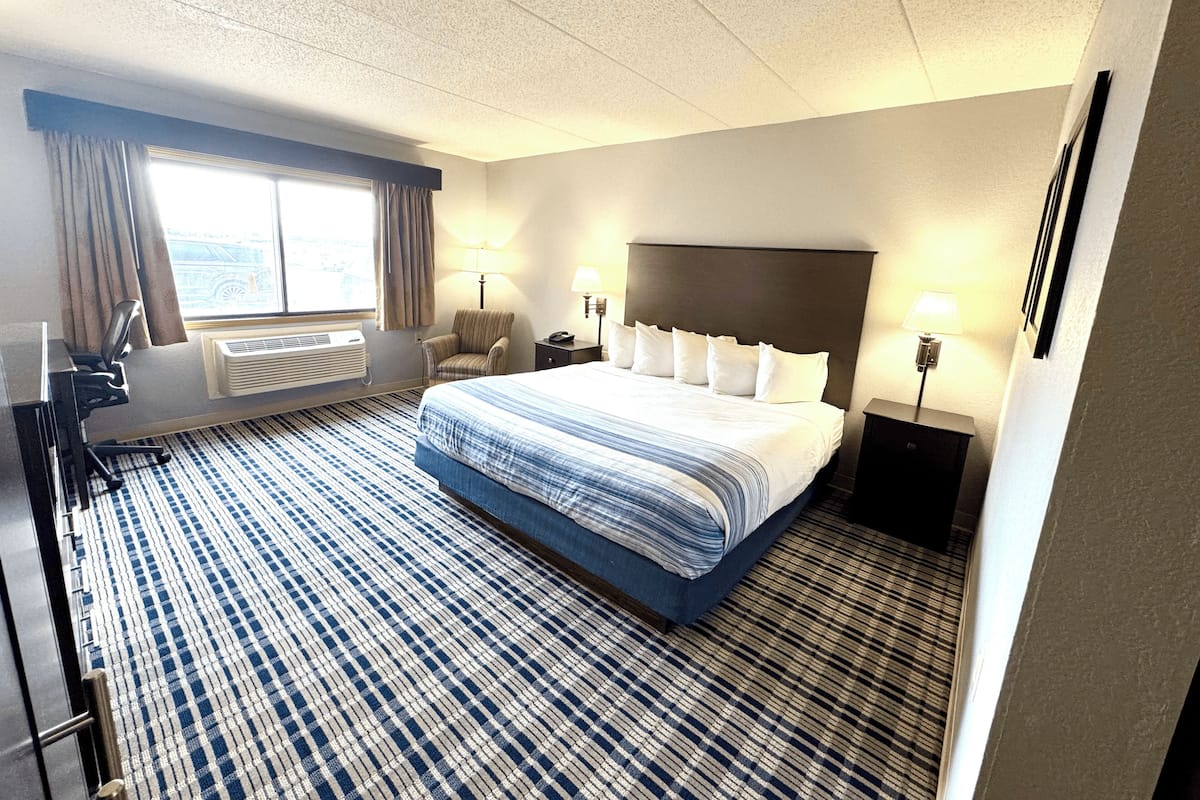 A hotel room with a large bed, two bedside tables with lamps, a desk with a chair, a window with curtains, and a radiator.